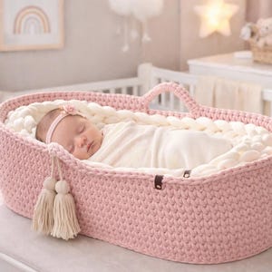 May include: A sleeping baby in a pink, hand-knitted bassinet. The oval-shaped bassinet has a handle and is filled with a soft, white blanket. The baby is wrapped in a white blanket and wearing a floral headband. Decorative tassels hang from the side.
