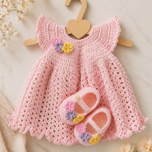 May include: A handmade, light pink crochet baby dress with ruffled sleeves and a scalloped hem, displayed on a wooden hanger. Matching crochet baby shoes with floral accents are placed below the dress. The dress features a small floral embellishment.