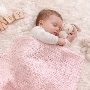 May include: A sleeping baby wrapped in a light pink crocheted blanket, clutching a stuffed bunny. The baby is wearing a white long-sleeved shirt. Wooden blocks spell out "BABY" in the background. A teddy bear is also visible.