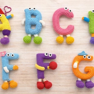 May include: A collection of colorful, crocheted letters of the alphabet, each with googly eyes, arms, and legs. The letters are in various colors, including yellow, blue, pink, green, purple, orange, and blue. The letters are arranged on a light wood surface.