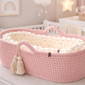 May include: A pink, oval-shaped baby bassinet with a thick, cream-colored interior. The bassinet has two handles and decorative tassels. The knitted design is visible, and the bassinet is placed on a white surface.