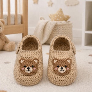 May include: A pair of beige crochet baby shoes with a bear face design on the front. The shoes have a rounded toe and a strap that fastens with a button. The background includes a teddy bear and a crib.