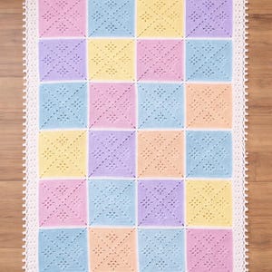 May include: A pastel-colored crochet blanket with a square pattern. The blanket features squares in pink, blue, yellow, and purple, each with a floral design. A white, scalloped border frames the blanket, which is displayed on a wooden surface.