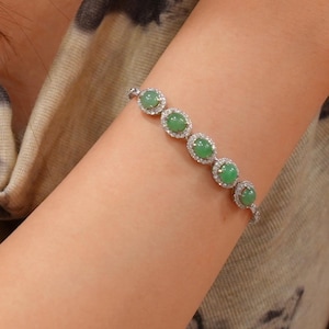 May include: A silver-toned bracelet featuring five oval-shaped, smooth green jade cabochons. Each jade stone is surrounded by a halo of small, clear, sparkling stones set in a metallic frame, connected by a delicate silver-toned chain link design.