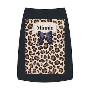 May include: Black dog shirt with a leopard print design. The shirt features a cream-colored oval with the name "Minnie" and a navy blue bow. The leopard print has brown and tan spots.