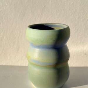 Puede incluir: Un jarrón de cerámica hecho a mano con una forma redondeada única. El jarrón presenta un degradado de colores, que va del verde suave en la base al azul pálido en la parte superior. El diseño es simple pero elegante, adecuado para exhibir flores o como pieza decorativa.