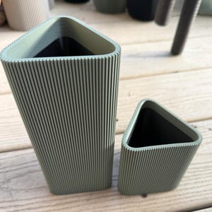 Puede incluir: Dos jardineras triangulares verde salvia con textura acanalada vertical. La jardinera más grande es más alta que la más pequeña. Ambas tienen un interior oscuro y se asientan sobre una superficie de madera. Las jardineras están vacías.