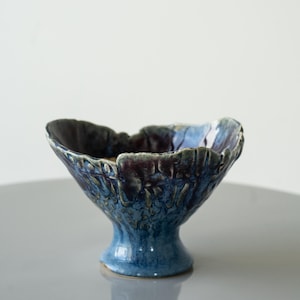 Puede incluir: Un cuenco de cerámica hecho a mano con una superficie texturizada única. El cuenco presenta una forma acampanada y orgánica con un borde azul oscuro y morado, que transiciona a una base azul más clara. El cuenco tiene aproximadamente 15 cm de diámetro.