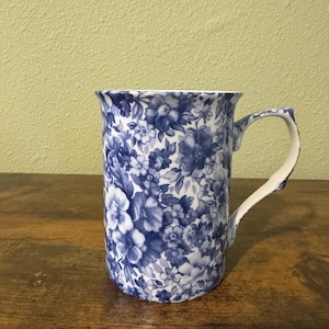Può includere: Una tazza in ceramica con manico bianco e motivo floreale blu. La tazza ha un bordo leggermente svasato ed è decorata con una fitta disposizione di fiori e foglie blu su sfondo bianco. La tazza è appoggiata su una superficie di legno.