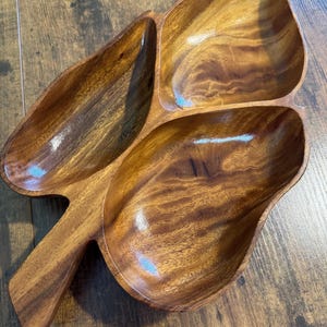 Puede incluir: Una bandeja de madera para servir con tres compartimentos en forma de hoja y un asa. La bandeja está hecha de madera de tonos cálidos con vetas visibles. Los compartimentos están diseñados para contener aperitivos.