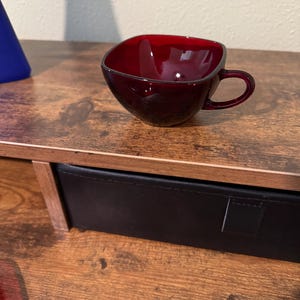 May include: A ruby red glass teacup with a square-shaped bowl and a rounded handle. The cup has a glossy finish and a dark rim. The teacup is sitting on a wooden surface.