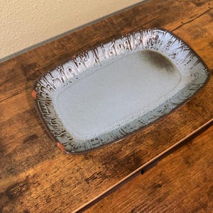 Peut inclure: Un plateau de service rectangulaire en céramique avec un intérieur gris clair et un bord brun foncé texturé. Le bord présente un motif détaillé avec de petits accents orange. Le plateau est présenté sur une surface en bois.