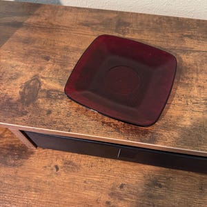 May include: A square, burgundy-colored glass plate with a circular indentation in the center. The plate is sitting on a wooden surface. The plate is a deep, transparent red.