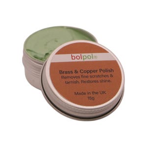 May include: A small metal tin of green polish with its lid beside it. The lid is brown with white text that reads "Brass & Copper Polish" and "Made in the UK 15g". The polish is designed to remove scratches and tarnish.
