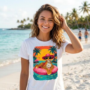 May include: White t-shirt featuring a colorful graphic of a leopard wearing sunglasses and a pink hat, relaxing on a pink inflatable ring in the ocean. The design includes palm trees, a sunset, and a flamingo.