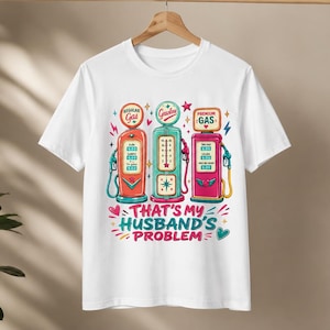 Pode incluir: T-shirt branca com um gráfico colorido de três bombas de gasolina retrô em vermelho, azul-petróleo e rosa. O texto "That's my husband's problem" está abaixo das bombas em uma fonte lúdica. A camisa está pendurada em um cabide de madeira.