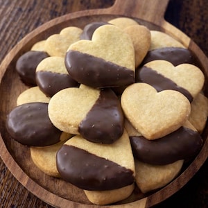 May include: A wooden serving platter piled high with heart-shaped biscuits. Each biscuit is partially dipped in dark chocolate. The biscuits are a light tan colour, and the chocolate is a rich brown. The platter has a handle.