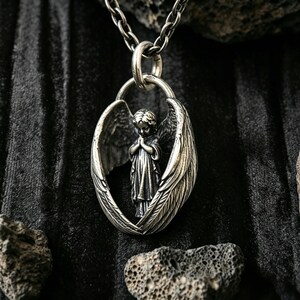 May include: A silver pendant necklace with a detailed angel figure, hands clasped in prayer, within textured wings. The pendant hangs from a matching chain. The background is dark and textured, with a focus on the intricate details of the jewellery.