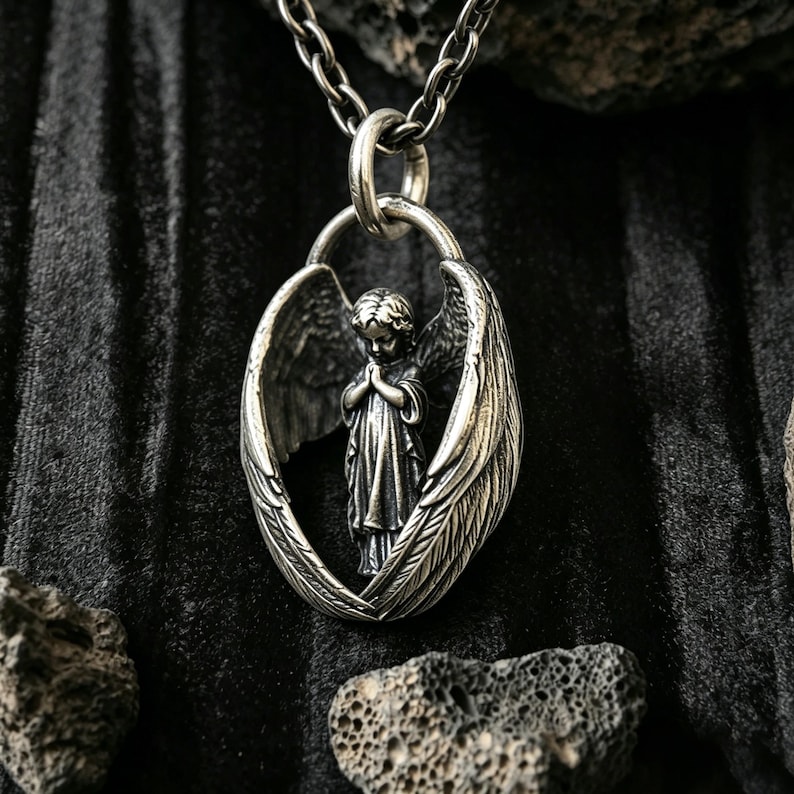 May include: A silver pendant necklace with a detailed angel figure, hands clasped in prayer, within textured wings. The pendant hangs from a matching chain. The background is dark and textured, with a focus on the intricate details of the jewellery.