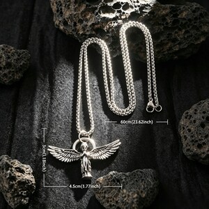 May include: Silver necklace with an angel pendant. The pendant is 4.5 cm wide and 3 cm tall. The chain is 60 cm long. The necklace is shown against a dark backdrop with rough, dark grey rocks, highlighting the jewellery's design.