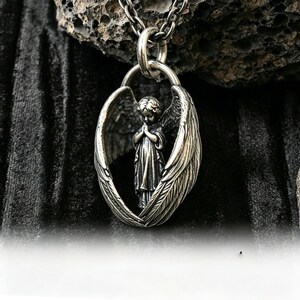 May include: A silver pendant necklace with a detailed angel figure framed by outstretched wings. The angel is shown with hands clasped in prayer. The necklace hangs from a silver chain, set against a dark, textured backdrop.