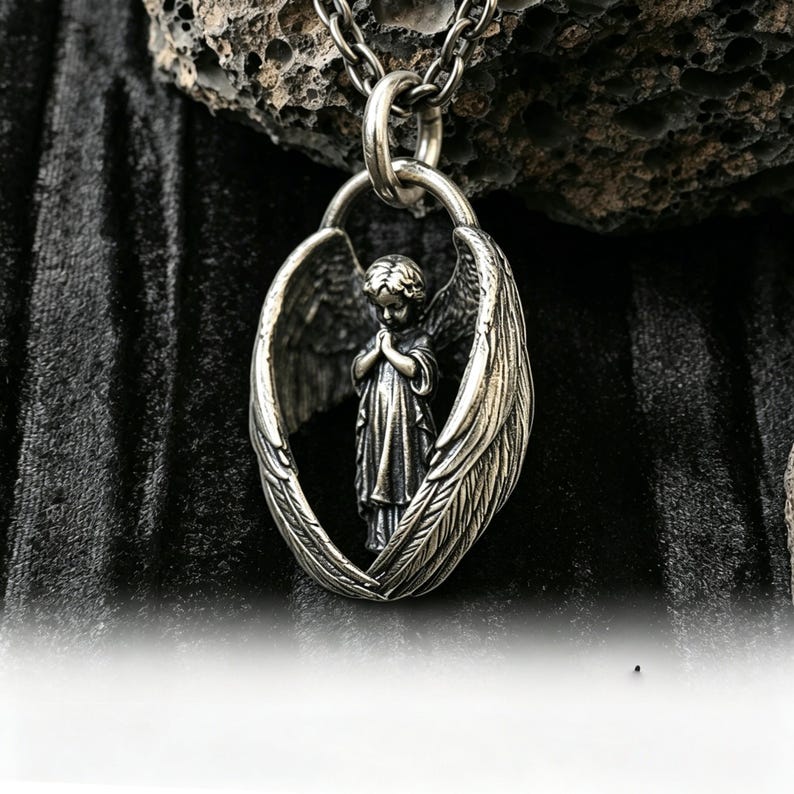 May include: A silver pendant necklace with a detailed angel figure framed by outstretched wings. The angel is shown with hands clasped in prayer. The necklace hangs from a silver chain, set against a dark, textured backdrop.