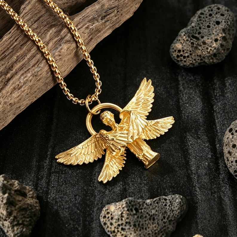 May include: A gold-coloured pendant necklace with an angel design. The pendant shows an angel with outstretched wings. The necklace chain is also gold-coloured. The background includes dark fabric and rough, dark grey stones.