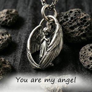 May include: A silver pendant necklace with an angel figure enclosed by wings. The angel is depicted with hands clasped in prayer. The words "You are my angel" are at the bottom. The necklace hangs on a chain.