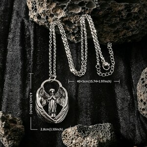May include: Silver pendant necklace with an angel figure and wings. The pendant is 4.3cm high and 2.8cm wide, suspended from a chain that is 40+5cm long. The necklace is displayed on a dark, textured surface. A detailed piece of jewellery.
