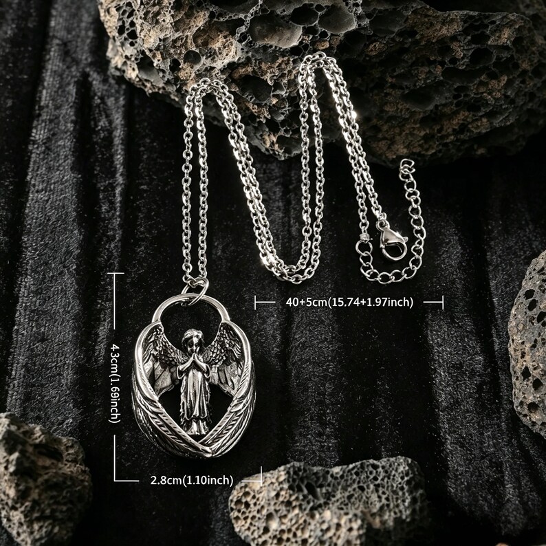 May include: Silver pendant necklace with an angel figure and wings. The pendant is 4.3cm high and 2.8cm wide, suspended from a chain that is 40+5cm long. The necklace is displayed on a dark, textured surface. A detailed piece of jewellery.
