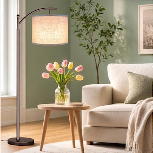 May include: A bronze-colored arc floor lamp with a beige drum shade illuminates a living room. A small wooden side table holds a vase of tulips. A cream-colored sofa and green pillow are in the background.