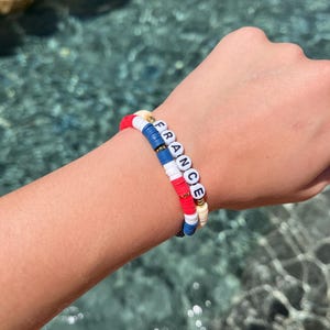 Puede incluir: Un conjunto de dos pulseras hechas de pequeñas cuentas de arcilla planas y circulares en rojo, blanco y azul. Una pulsera presenta cuentas de letras blancas que deletrean la palabra FRANCE en negro, con pequeñas cuentas espaciadoras metálicas doradas.