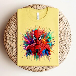 Op de afbeelding: Geel T-shirt met een afbeelding van Spider-Man. De superheld is afgebeeld in een rood en zwart pak, tegen een achtergrond van kleurrijke verfspatten. Het shirt is op een geweven oppervlak gevouwen.