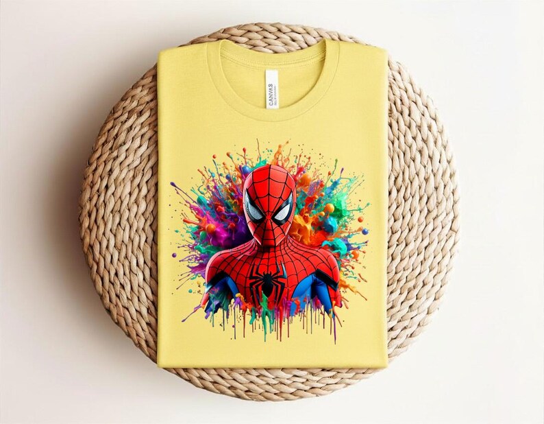 Op de afbeelding: Geel T-shirt met een afbeelding van Spider-Man. De superheld is afgebeeld in een rood en zwart pak, tegen een achtergrond van kleurrijke verfspatten. Het shirt is op een geweven oppervlak gevouwen.