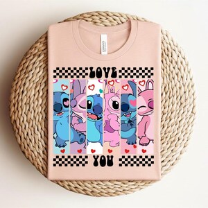 May include: A light pink t-shirt featuring a graphic of cartoon characters with the words "LOVE YOU" in black. The graphic includes blue, pink, and purple characters with hearts and a checkered pattern.