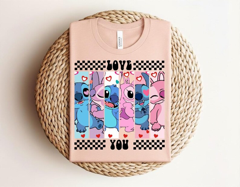 May include: A light pink t-shirt featuring a graphic of cartoon characters with the words "LOVE YOU" in black. The graphic includes blue, pink, and purple characters with hearts and a checkered pattern.