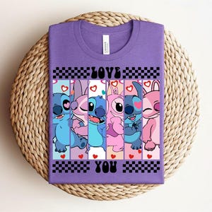 May include: A purple t-shirt featuring a graphic design with the text "LOVE YOU" and cartoon characters. The design includes blue, pink, and purple characters with hearts and checkered patterns.