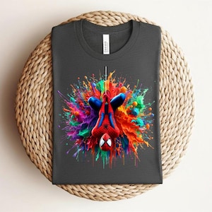 May include: Dark gray t-shirt featuring a vibrant, colorful graphic of Spider-Man hanging upside down. The design showcases Spider-Man in his classic red and blue suit, set against a splash of rainbow colors. The shirt is displayed on a woven surface.
