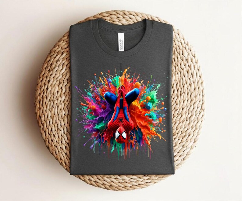 May include: Dark gray t-shirt featuring a vibrant, colorful graphic of Spider-Man hanging upside down. The design showcases Spider-Man in his classic red and blue suit, set against a splash of rainbow colors. The shirt is displayed on a woven surface.