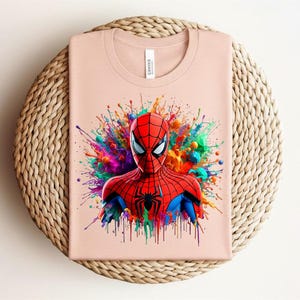 Op de afbeelding: Een lichtroze t-shirt met een kleurrijke afbeelding van Spider-Man. De superheld is afgebeeld in een rood en zwart kostuum, tegen een levendige splash van regenboogkleuren. Het shirt is opgevouwen op een geweven, cirkelvormige mat.