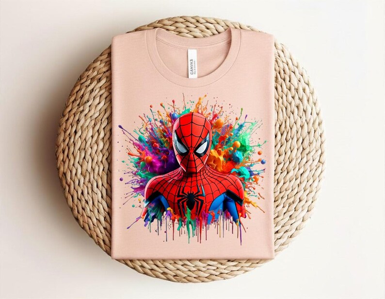 Op de afbeelding: Een lichtroze t-shirt met een kleurrijke afbeelding van Spider-Man. De superheld is afgebeeld in een rood en zwart kostuum, tegen een levendige splash van regenboogkleuren. Het shirt is opgevouwen op een geweven, cirkelvormige mat.
