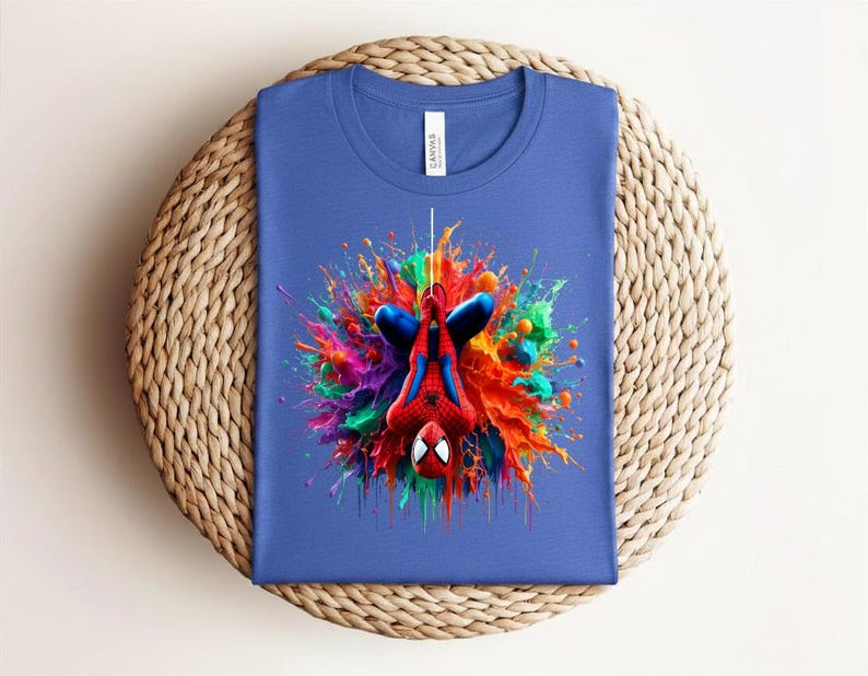 May include: Blue t-shirt featuring a colorful graphic of Spider-Man hanging upside down. The design includes vibrant paint splatters in red, orange, green, and purple. The shirt is made of a soft material and is suitable for casual wear.