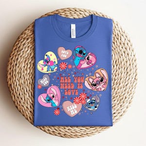 Peut inclure: Un t-shirt bleu avec un motif color&eacute; de personnages de dessins anim&eacute;s en forme de c&oelig;ur. Le motif comprend le texte "ALL YOU NEED IS LOVE" et des phrases comme "LOVE YOU" et "KISS ME".