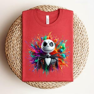 May include: A red crew neck t-shirt featuring a graphic of a white skull character with a stitched mouth and black suit, centered against a vibrant, multi-colored paint splatter design with splashes of blue, green, orange, and yellow hues.