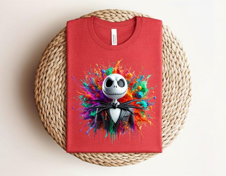 May include: A red crew neck t-shirt featuring a graphic of a white skull character with a stitched mouth and black suit, centered against a vibrant, multi-colored paint splatter design with splashes of blue, green, orange, and yellow hues.