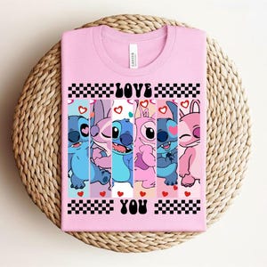 May include: A light pink t-shirt featuring a graphic of cartoon characters in shades of blue, pink, and purple. The words "LOVE YOU" are printed in black, with a checkered pattern above and below the text. Hearts are scattered throughout the design.