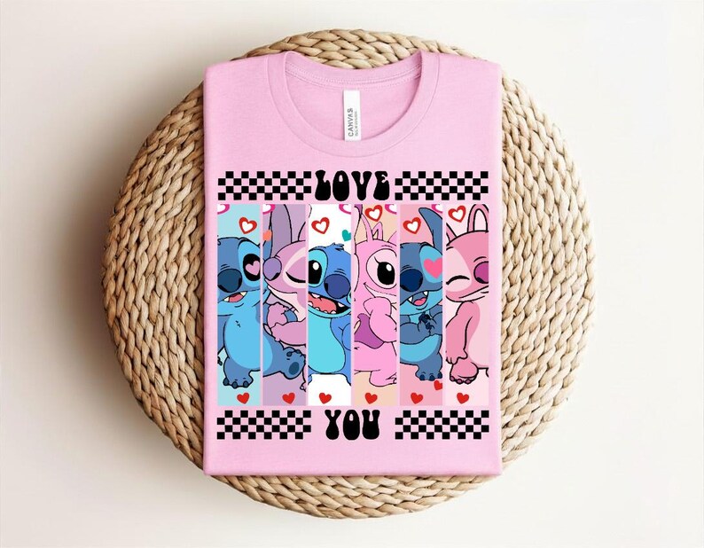 May include: A light pink t-shirt featuring a graphic of cartoon characters in shades of blue, pink, and purple. The words "LOVE YOU" are printed in black, with a checkered pattern above and below the text. Hearts are scattered throughout the design.