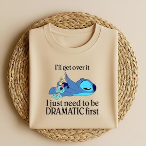 May include: A beige t-shirt featuring a graphic of the character Stitch from the movie Lilo & Stitch. Stitch is depicted in blue, lying down with a sad expression. The text "I'll get over it, I just need to be DRAMATIC first" is printed on the shirt.