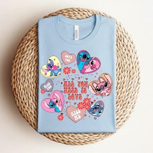 Peut inclure: T-shirt bleu clair avec des personnages de dessins anim&eacute;s en forme de c&oelig;ur. Le t-shirt porte l'inscription "All You Need Is Love" et d'autres phrases. Le t-shirt est pli&eacute; sur une surface tiss&eacute;e.