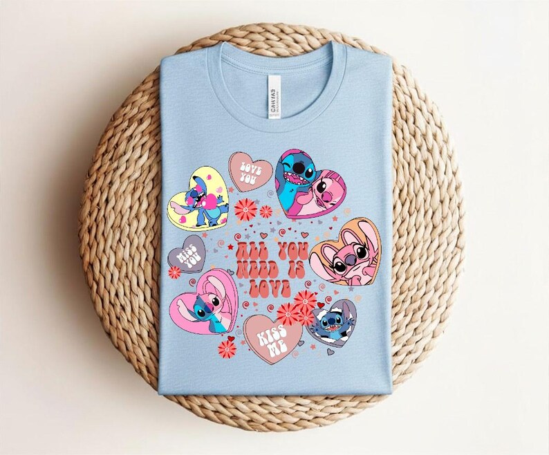 Peut inclure: T-shirt bleu clair avec des personnages de dessins anim&eacute;s en forme de c&oelig;ur. Le t-shirt porte l'inscription "All You Need Is Love" et d'autres phrases. Le t-shirt est pli&eacute; sur une surface tiss&eacute;e.